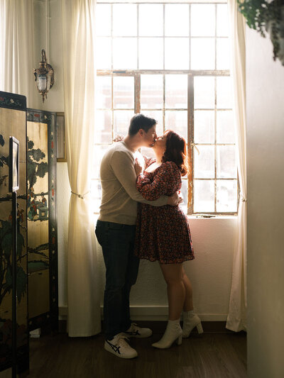 A couple kisses in front of a window in their favorite local wine bar during their engagement portraits.