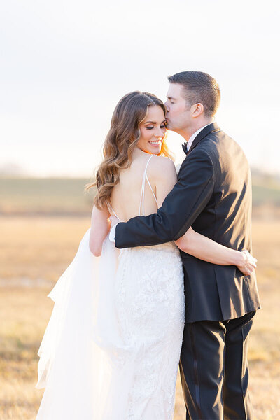 Bride and groom in a field with sun setting be3hind them. Groom is kissing the forehead of his bride.