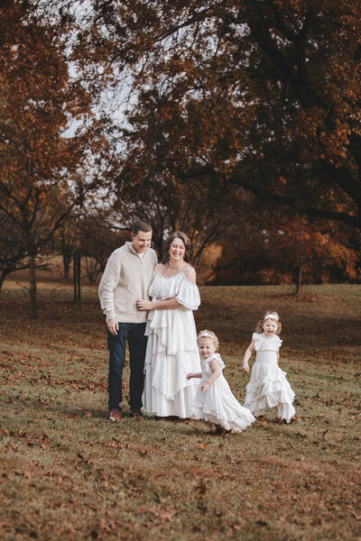 Family Portraits of a young family parents in the middle embracing each other with toddler girls running circles around them having fun