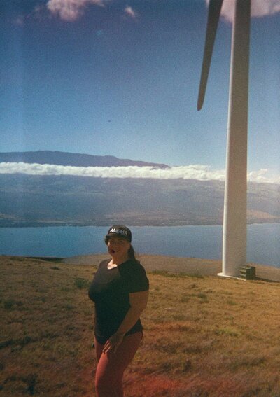 photo of Mariah with a hat in front of a wind turbine, there are sea and land in the distance on Maui