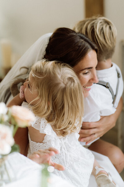 Bruid met kinderen tijdens haar bruiloft, vastgelegd in een ongedwongen en avontuurlijke sfeer, waar plezier en spontaniteit centraal staan. Knuffel van moeder met kinderen. 