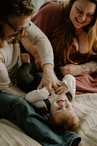 Family photo of mom and dad on bed wearing cream and mauve while dad is tickling baby in studio in Baltimore Maryland.