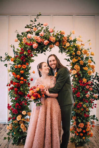Couple stands under rainbow floral arch