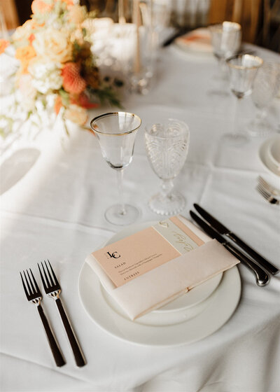 Peach wedding menu with custom monogram tucked into a napkin on a wedding table with floral details and flatware representing full service design and calligraphy by Life in Letters.