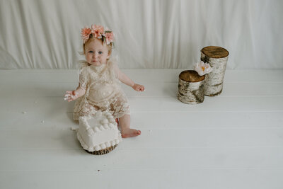 Baby girl with cake on her hand while sitting behind a cake and wearing flower crown with white lace dress in Annapolis Maryalnd.