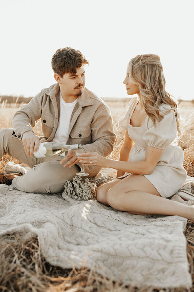 couple sits on blanket in field while the man pours a glass of champagne