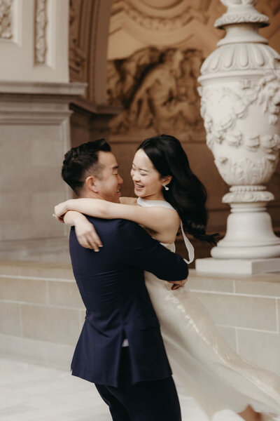 A joyful couple embracing at San Francisco City Hall, with the man lifting the woman slightly off the ground, both smiling, in an elegant setting with classical architecture.