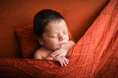 new born baby with dark hair laying under orange blanket
