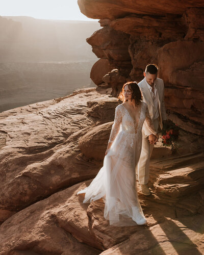 bride and groom standing on the side of a cliff with the desert below them