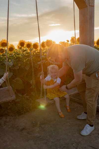 toddler girl smiles as father sets her on wooden swing in the middle of a sunflower field at gust flower and produce farm in ottawa lake michigan