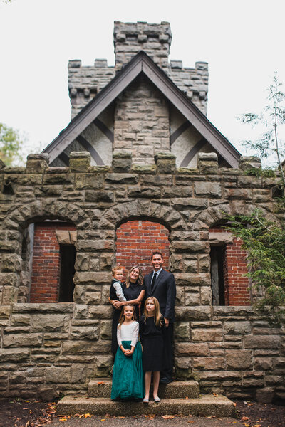 Family photographer capturing a mom, dad, and three children outside next to a stone building.