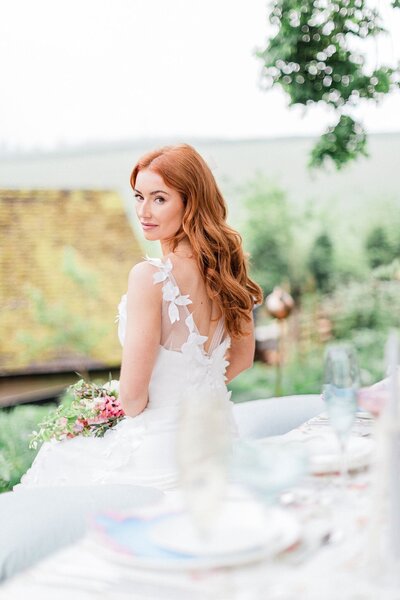Bride with long red hair in a floral appliqué gown looking over her shoulder during a romantic outdoor wedding setup in the English countryside