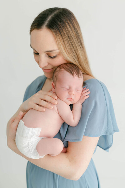 New Mom in a blue chiffon dress holding newborn baby up against chest with baby's head facing the camera. Mom is resting her cheek on the back of baby's head and looking down and smiling.