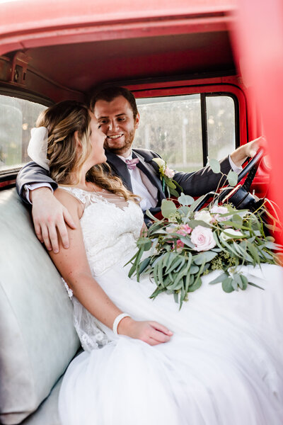 Bride in white lace dress holding flowers on her lap leaned against groom as he looks at her sitting in red vintage truck