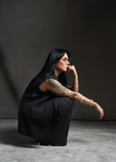 Person with long dark hair, wearing glasses and black clothing, crouches on the floor with their hand on their chin. Showcasing visible tattoos and multiple bracelets, they exude a creative aura that speaks to the meticulousness required to hire an online business manager.