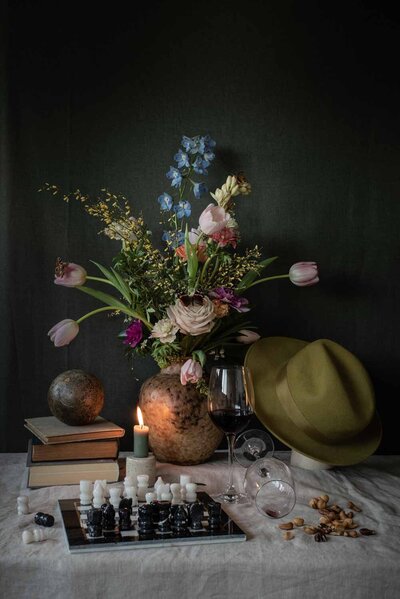 Stilleven met pastelkleurig bloemstuk met diverse vlinders in vaas van aardewerk, met rondom vintage boeken, hoed, schaakspel en glas met rode wijn.