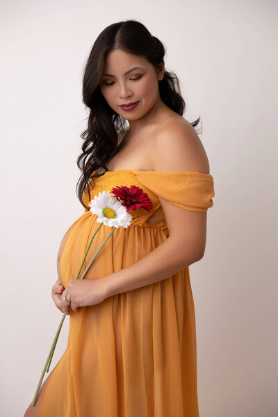 Maternity photo of expecting mom wearing a yellow off-the-shoulder dress and looking down while holding a white and red daisy flower.