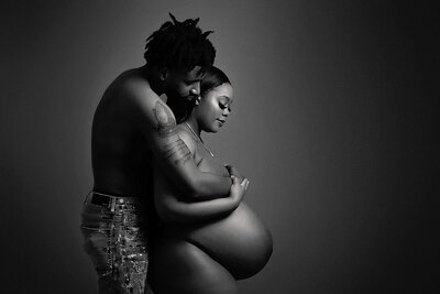 A black and white photo by a Jacksonville maternity photographer shows a couple embracing. The pregnant woman, partially nude, stands in front, with her partner, shirtless and in jeans, standing behind her and wrapping his arms around her in a tender hold. The background is plain and dark.