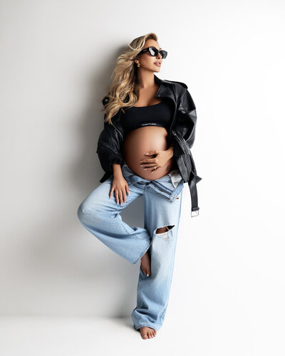 A pregnant woman in leather jacket and jeans against a neutral backdrop, showcasing modern maternity photography.