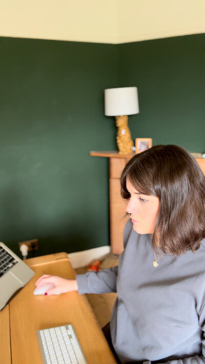 a female social media manager sits at an oak table. she is using a mouse to work her laptop. 