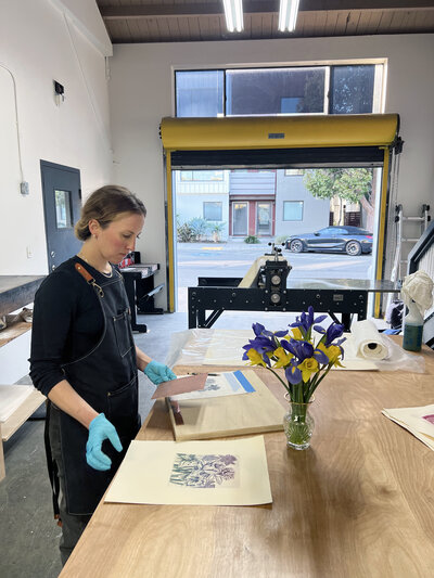 A focused printmaker examines a freshly pressed print beside a vibrant vase of purple and yellow flowers in a sunny printmaking studio.