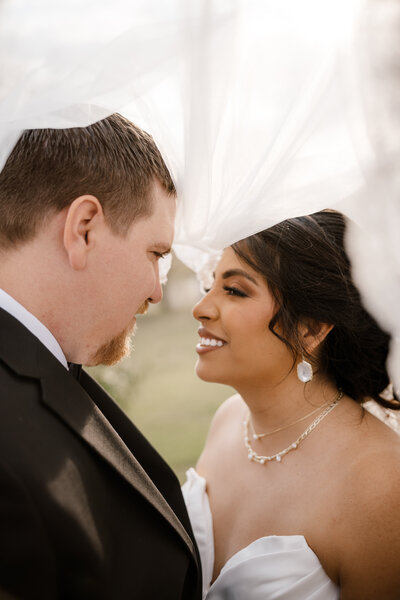 Bride and groom take a picture under the brides veil smiling at each other while being captured by Shelly Voss