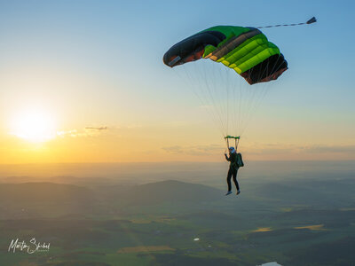 Das Bild zeigt einen Fallschirmspringer, der die Abendstimmung unter seinem Schirm genießt. Zu sehen sind die Berge im Hintergrund und der Sonnenuntergang