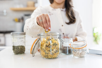 Close up of a woman's hands reaching into a jar with dried flowers and herbs for her Ayurvedic practice.