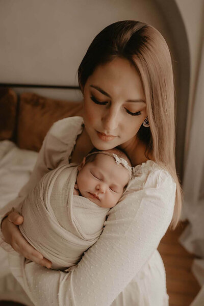 A mother embracing her new baby girl in a cream swaddle and floral headband in a gorgeous natural light  studio in Canandaigua, NY