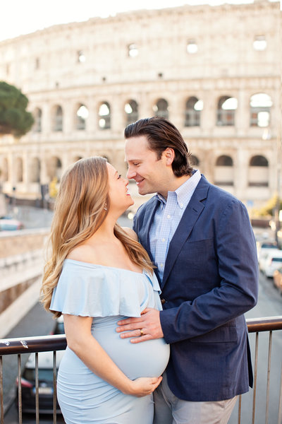 A pregnant woman and her husband with hands on her belly and looking into each others eyes in front of the Colosseum. Taken by Rome Photographer, Tricia Anne Photography.