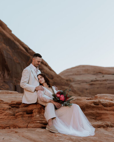 bride and groom sitting on the ground in the desert with the brides face leaned into the grooms chest