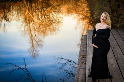 Stunning outdoor maternity photography featuring an expectant mother posed gracefully on a pier, with a serene water reflection showcasing the golden hour light and surrounding nature