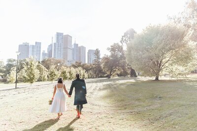 A bride and groom walking through a park with the Melbourne CBD buildings in the backdrop.