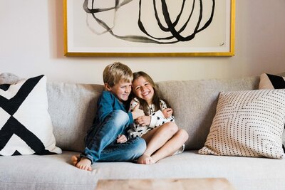 Two children sitting on a couch, smiling and embracing, with a modern art piece hanging on the wall behind them in Pittsburgh.