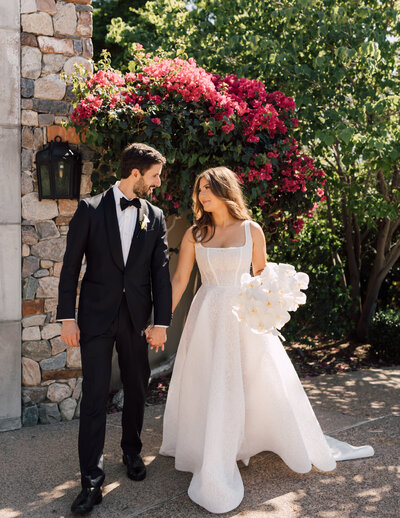 Bride and groom walking hand-in-hand in front of vibrant bougainvillea, captured by a Scottsdale wedding photographer in a scenic outdoor wedding venue.