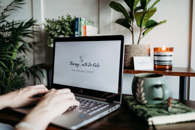 A cozy online therapy setup with a laptop, candle, and greenery beside a couch
