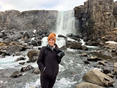 A photographer stands smiling at the camera with a camera in her hand. She is standing in front of a waterfall with water flowing over rocks all around her.