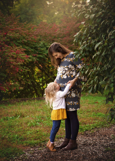Mom smiling down at young daughter kissing her pregnant belly.