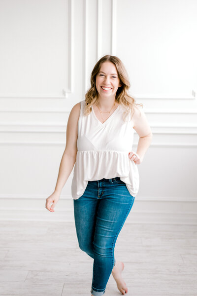 A headshot of Cleveland Family & Newborn Photographer, Brittany Serowski Photography owner, Brittany Serowski. Standing in front of an all white wall at the Lake Arts Studio in Avon Lake, OH, she is wearing dark blue jeans and a white tank top as she smiles walking towards the camera.