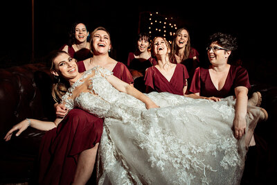 Bride takes photo in the arms of her bridesmaids wearing maroon dresses.