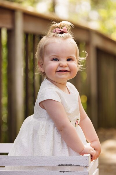 A baby girl in a white dress sits on a wooden bench, smiling and enjoying a sunny day outdoors.