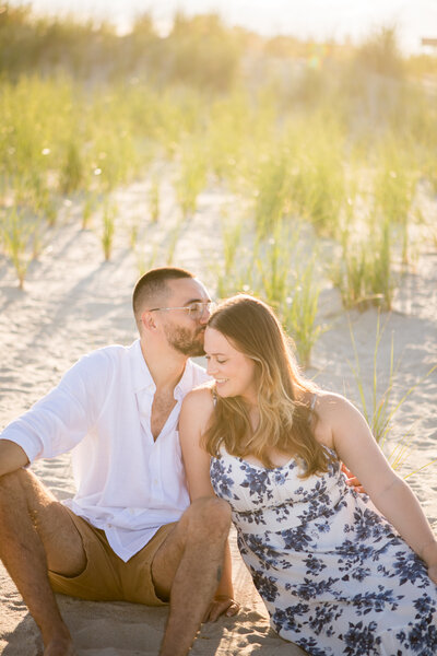 a couple sits in the dunes with hazy golden light behind them a few moments after they got engaged on long beach island 