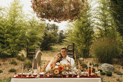 happy young couple just married, seated at their table, decorated in deep reds and oranges, bride rests her head on the grooms shoulder