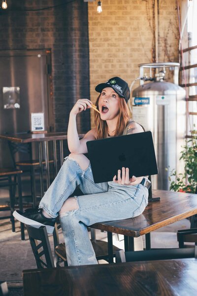 A woman with long red hair sits barefoot on a wooden block, wearing a black top and patterned pants, looking at the camera—the owner of an agency offering branding and web design for female entrepreneurs.