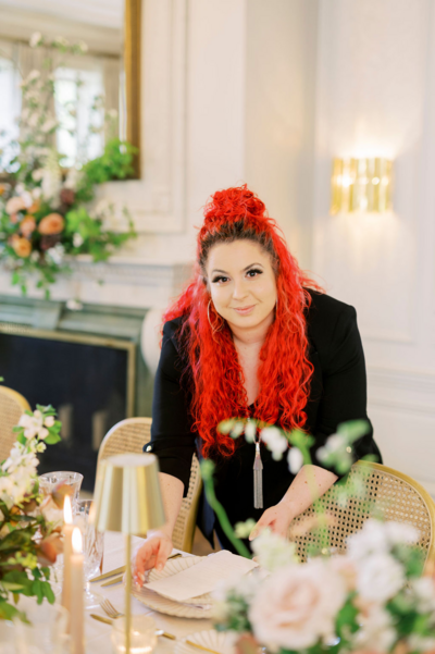 woman with bright red crimson hair smiling at the camera while adjusting a menu at table filled with soft blush florals and candles
