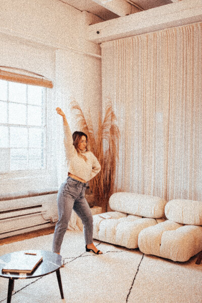 Girl is dancing in a living room while wearing a turtleneck, blue jeans and black heels