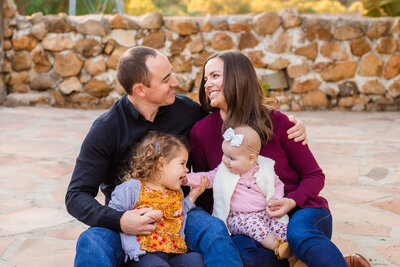 Family photographer, a family of 4 sit on the ground laughing while their two baby girls touch