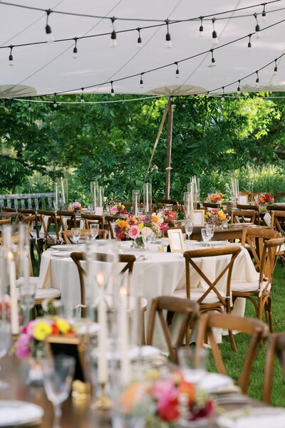 Elegant outdoor wedding reception setup under a tent with string lights, featuring round tables adorned with floral centerpieces and wooden chairs.