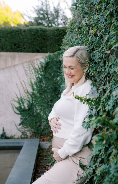 Mom holding her baby bump while leaning against a vine-covered wall.