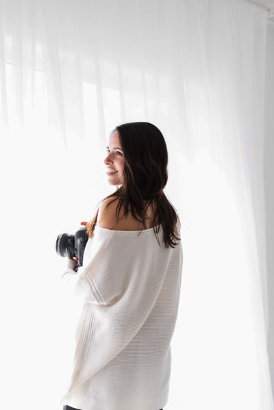 An image of Laurie Baker, photographer at Boudoir by Elle, headshot image of  a photographer in white sweater holding camera